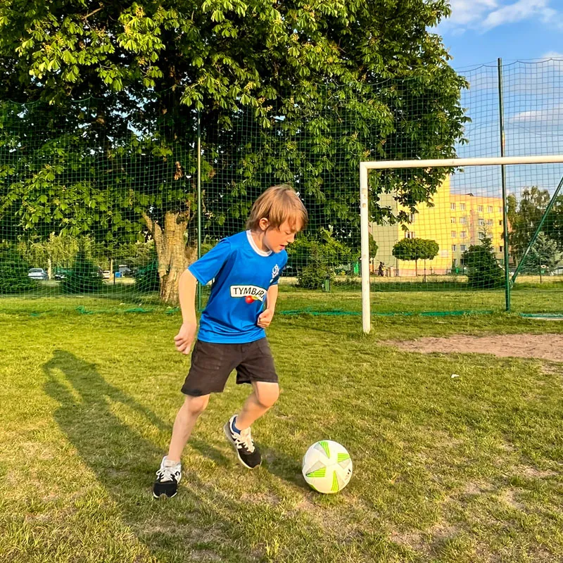 Chłopiec w niebieskiej koszulce piłkarskiej prowadzi piłkę na trawiastym boisku